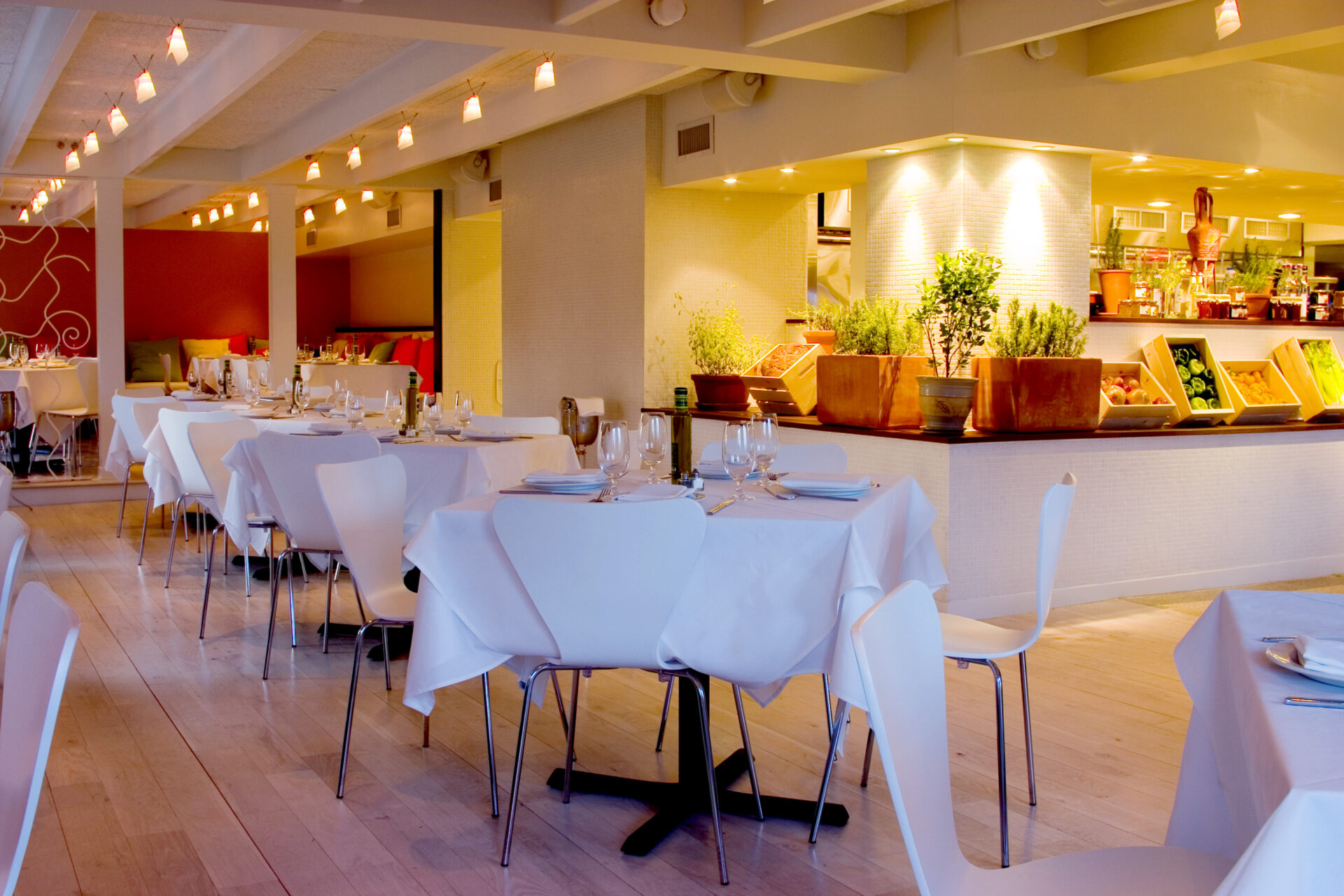 Intimate corner of Trata Restaurant featuring two-top tables with white cloths, colorful pillows on the bench, and a warm orange accent wall.