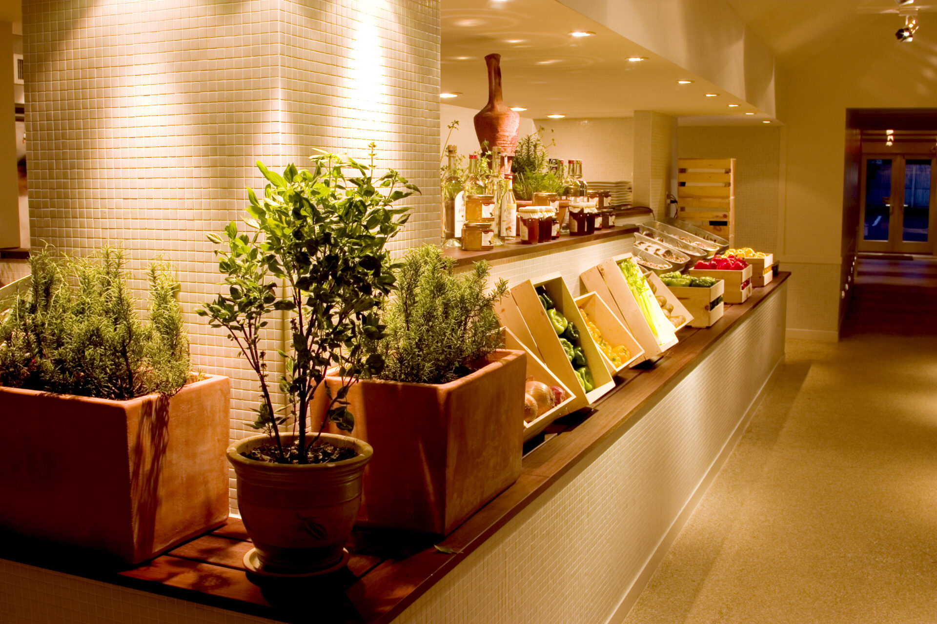 Close‑up of Trata’s specialty products on display, including jars of preserves, bottles of ouzo, olive oil, and potted herbs arranged beneath warm lighting.