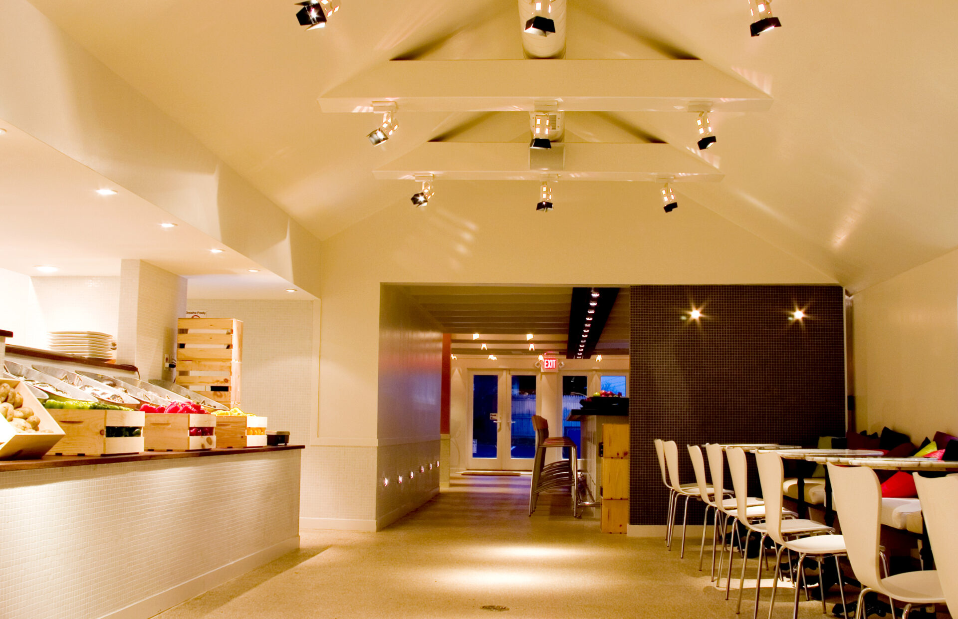 Entry axis of Trata Restaurant, with open kitchen and market‑style produce display on one side and a row of white café tables and chairs on the other.