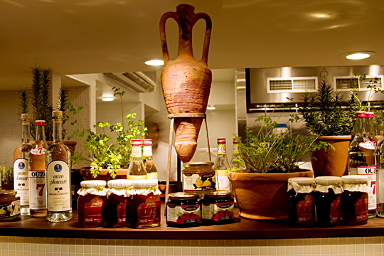 Angled shot of the open kitchen and market display at Trata Restaurant, with crates of vegetables and rows of potted herbs under spotlights.