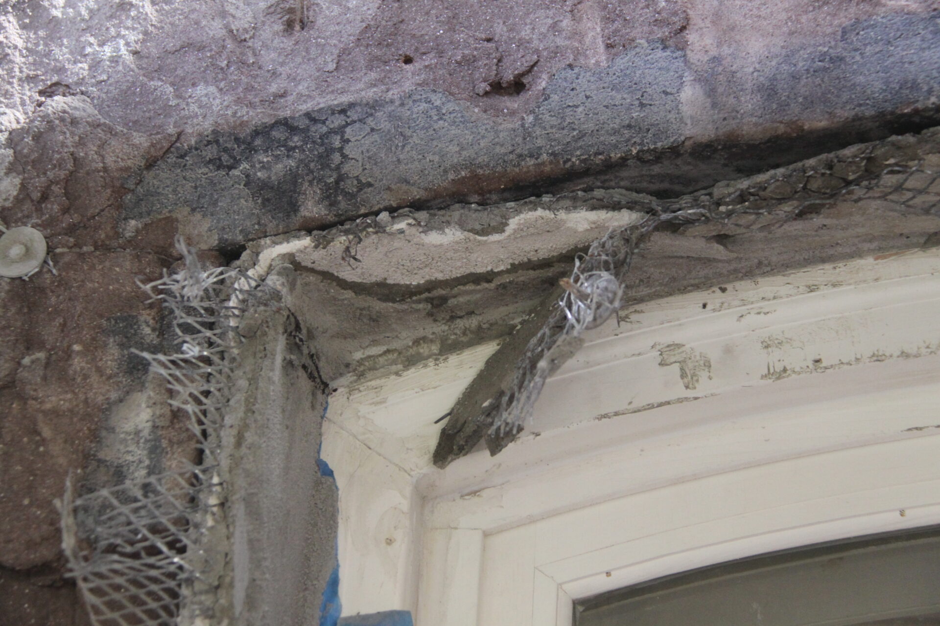Construction close-up revealing damaged historic stucco and lintel at Kim Eng Townhouse, with exposed metal lath and layers of deteriorated masonry above a window opening.