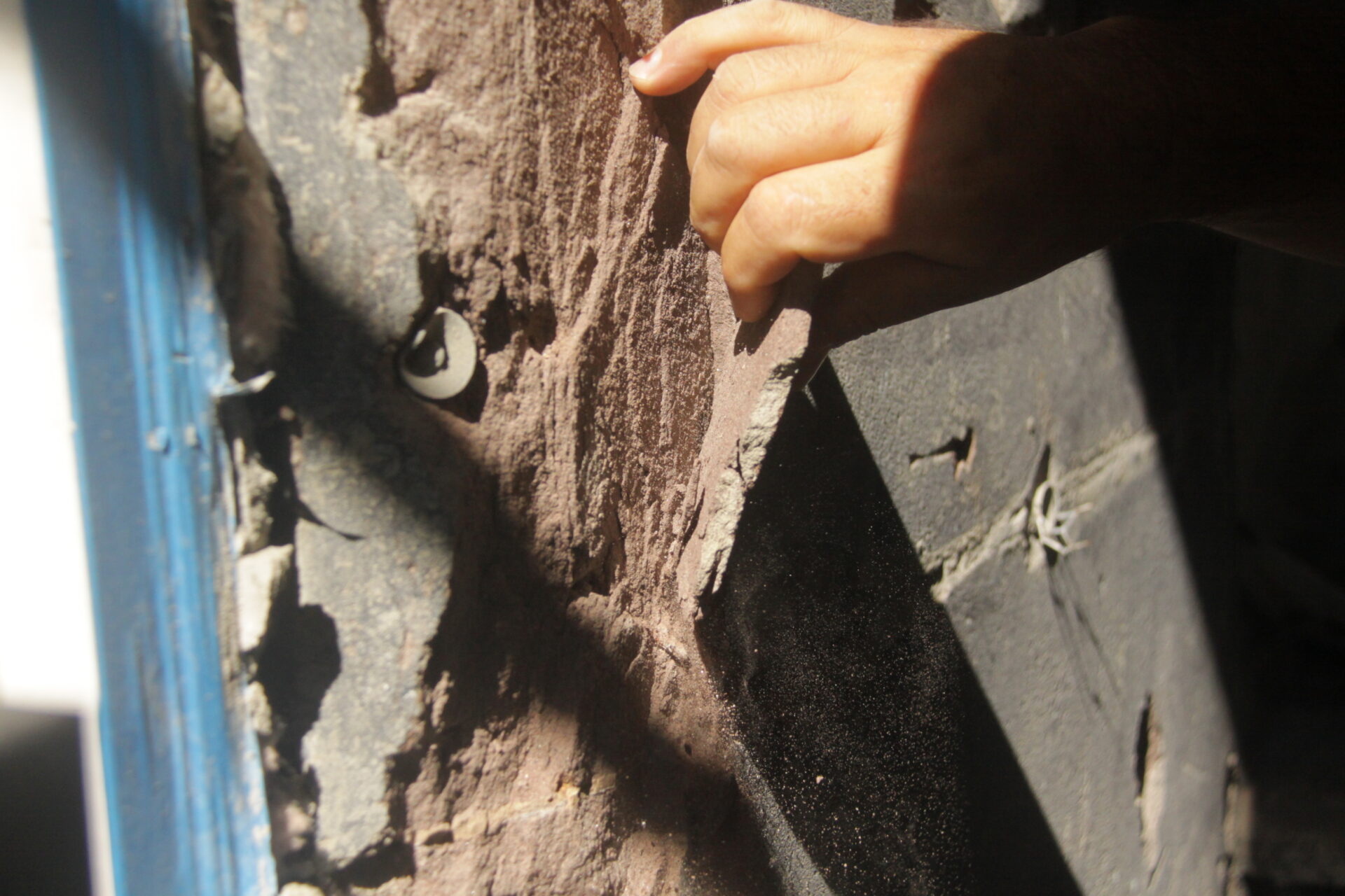 Detail of investigative work at Kim Eng Townhouse, showing a hand peeling back failed brownstone stucco to expose the substrate and anchoring system behind.