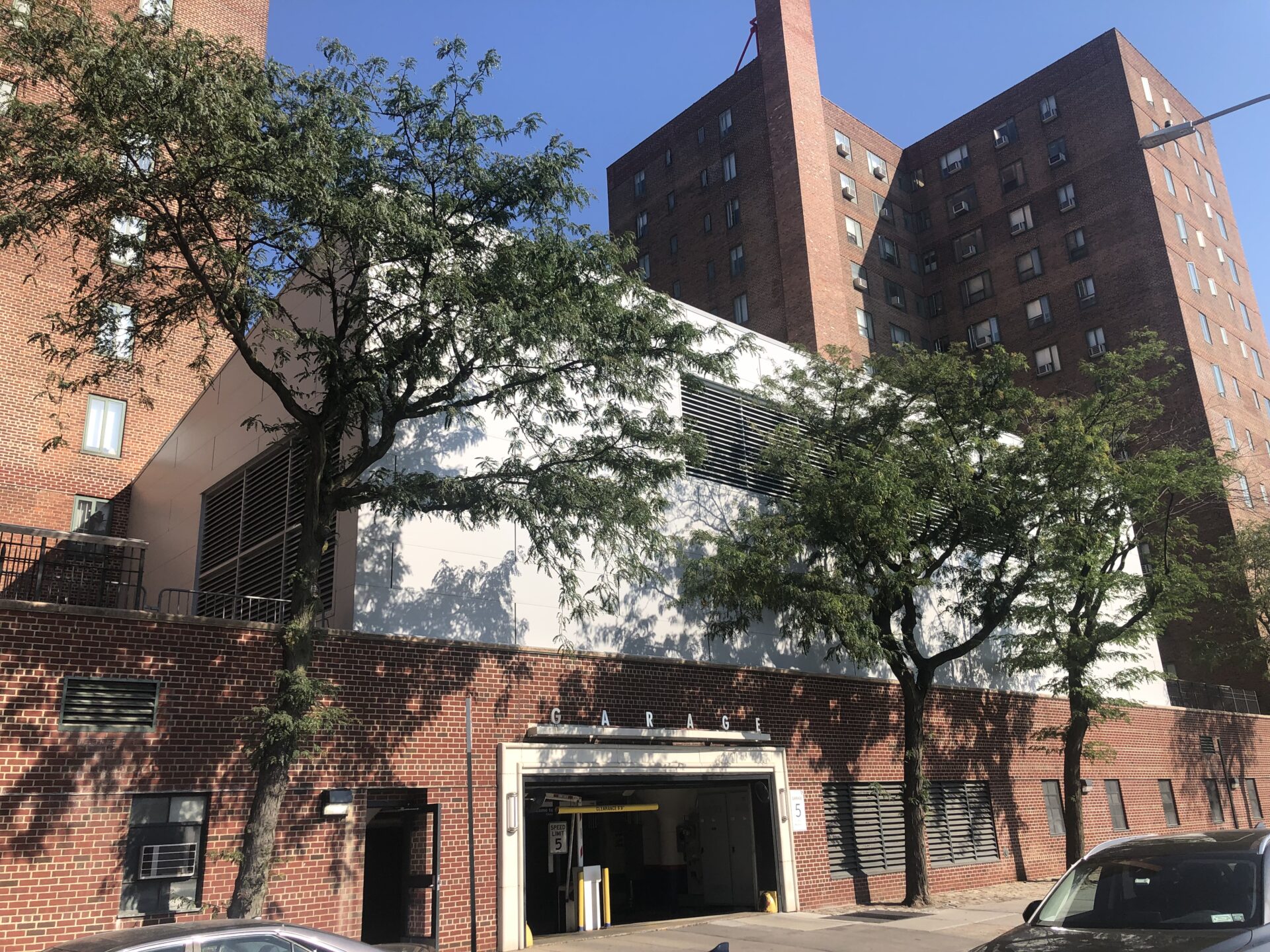 Angled street view of the energy building with its white panelized façade hovering above the brick garage base, framed by trees and the surrounding high-rise apartments.