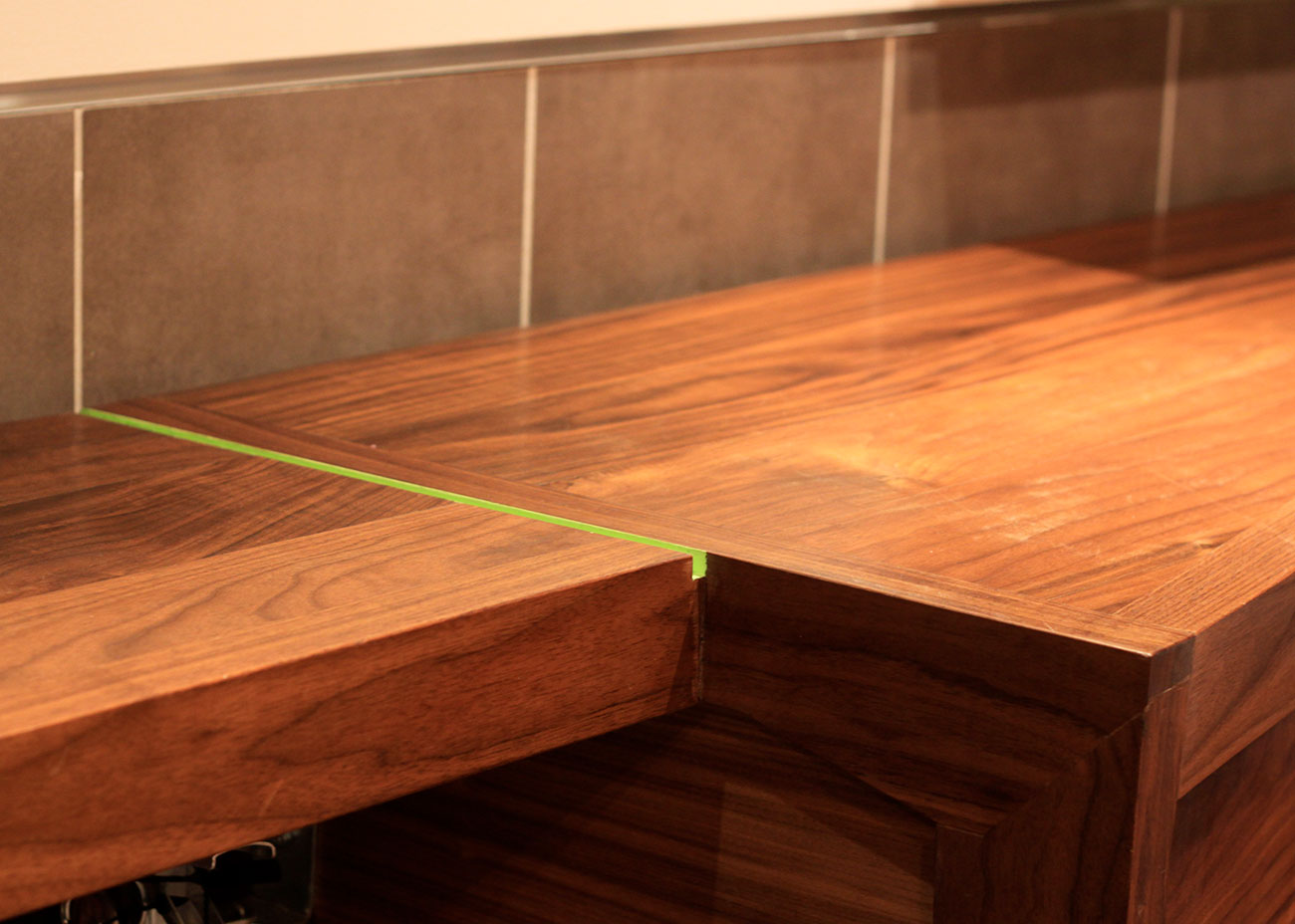Close-up of Kalamaki’s walnut wall counter showing the mitered corner joint and a thin inlay of bright green acrylic, set against a backdrop of gray rectangular wall tiles.