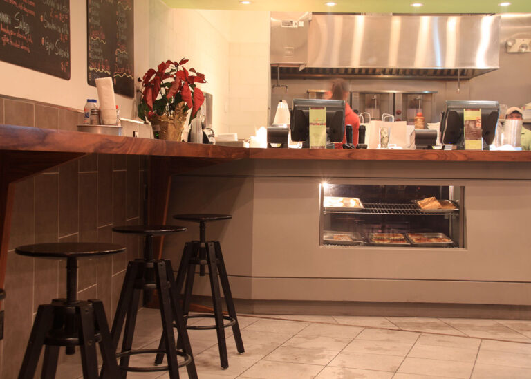 Lower portion of Kalamaki’s front counter with walnut top, display window of prepared dishes, and bar stools along the adjacent wall counter.