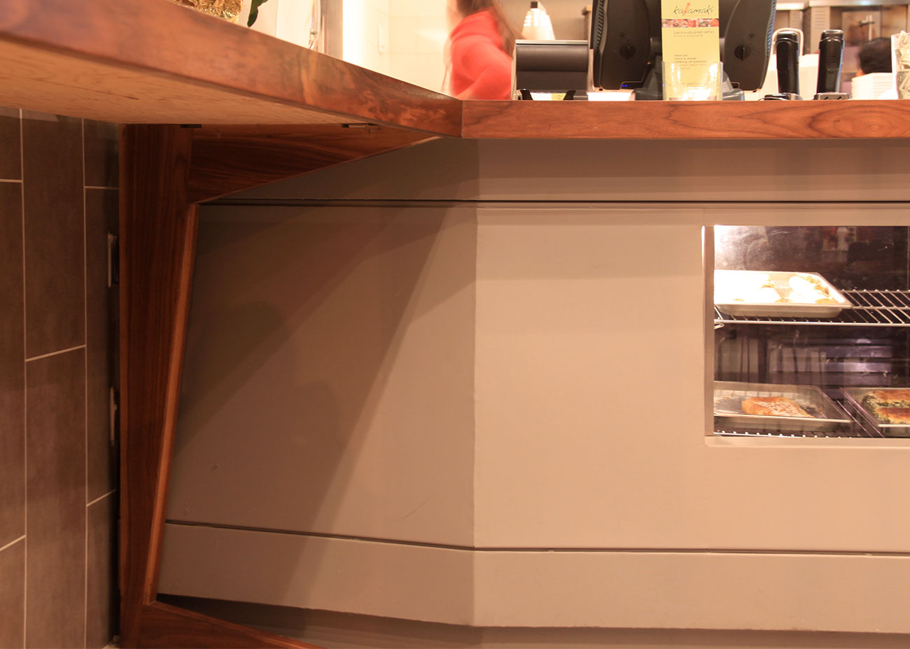 Detail of Kalamaki’s front counter showing the angled walnut support leg, gray counter base, and a lower display window with trays of food, with the cashier and open kitchen softly blurred in the background.