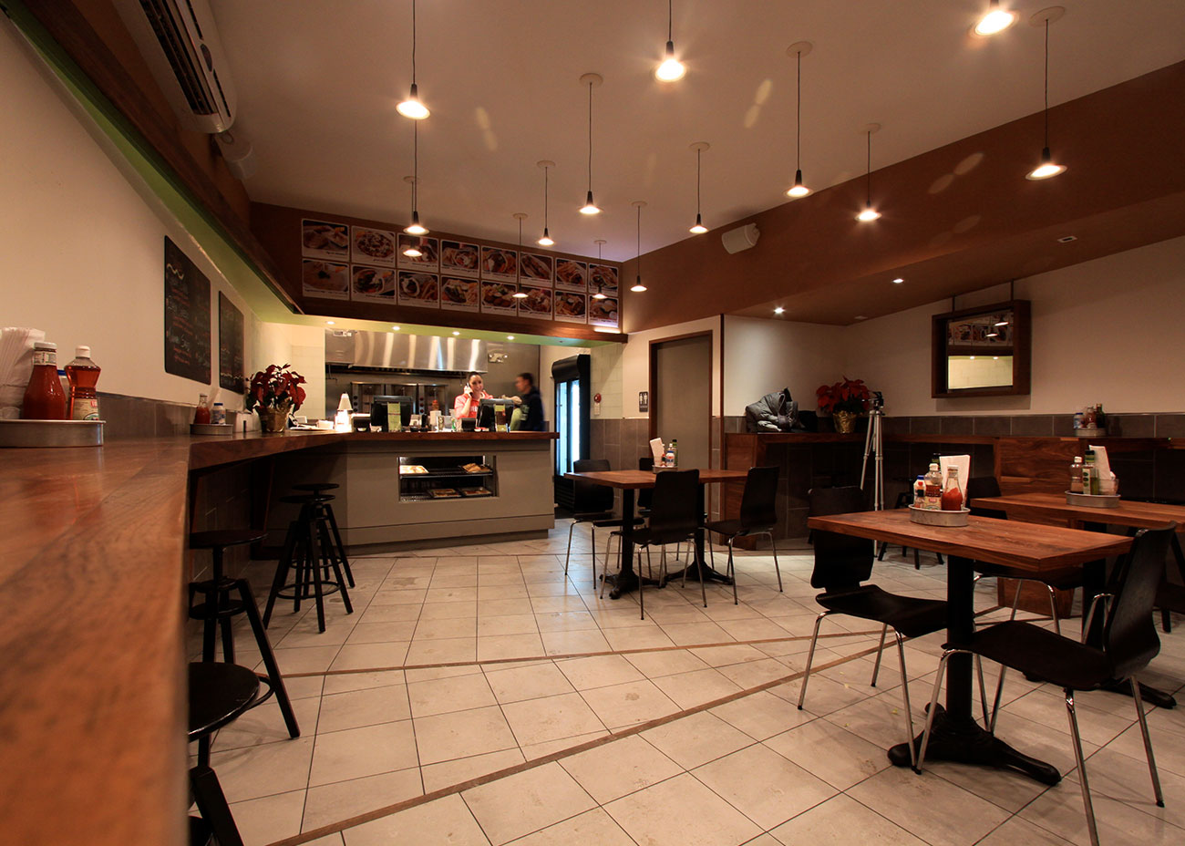 Seating at Kalamaki looking toward the ordering counter, with square walnut tables, black chairs, wall-hung mirrors, and the skewed ceiling band emphasizing the depth of the room.