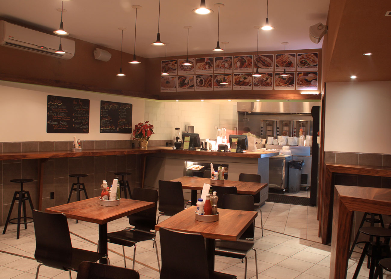 View from the window side of Kalamaki back to the ordering area, showing perimeter bar seating, freestanding tables, and warm walnut paneling framing the compact space.