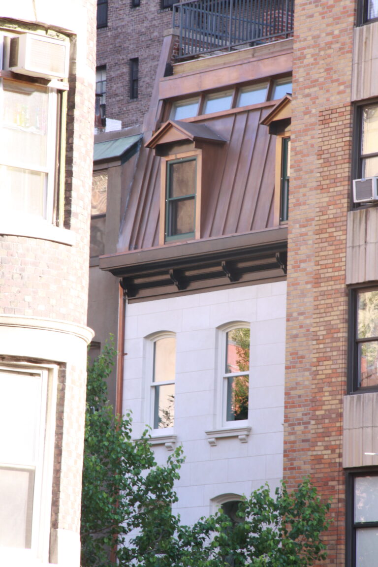 Distant streetscape view with Kim Eng Townhouse’s copper roofline and light façade emerging above and between mature trees.