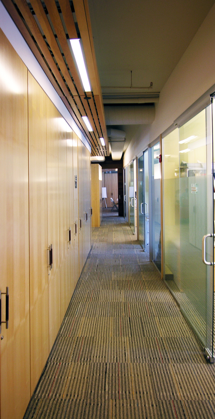Office corridor at Spence-Chapin lined with full-height maple storage cabinets on one side and glass-fronted meeting rooms on the other, with linear lights set into a slatted wood ceiling.