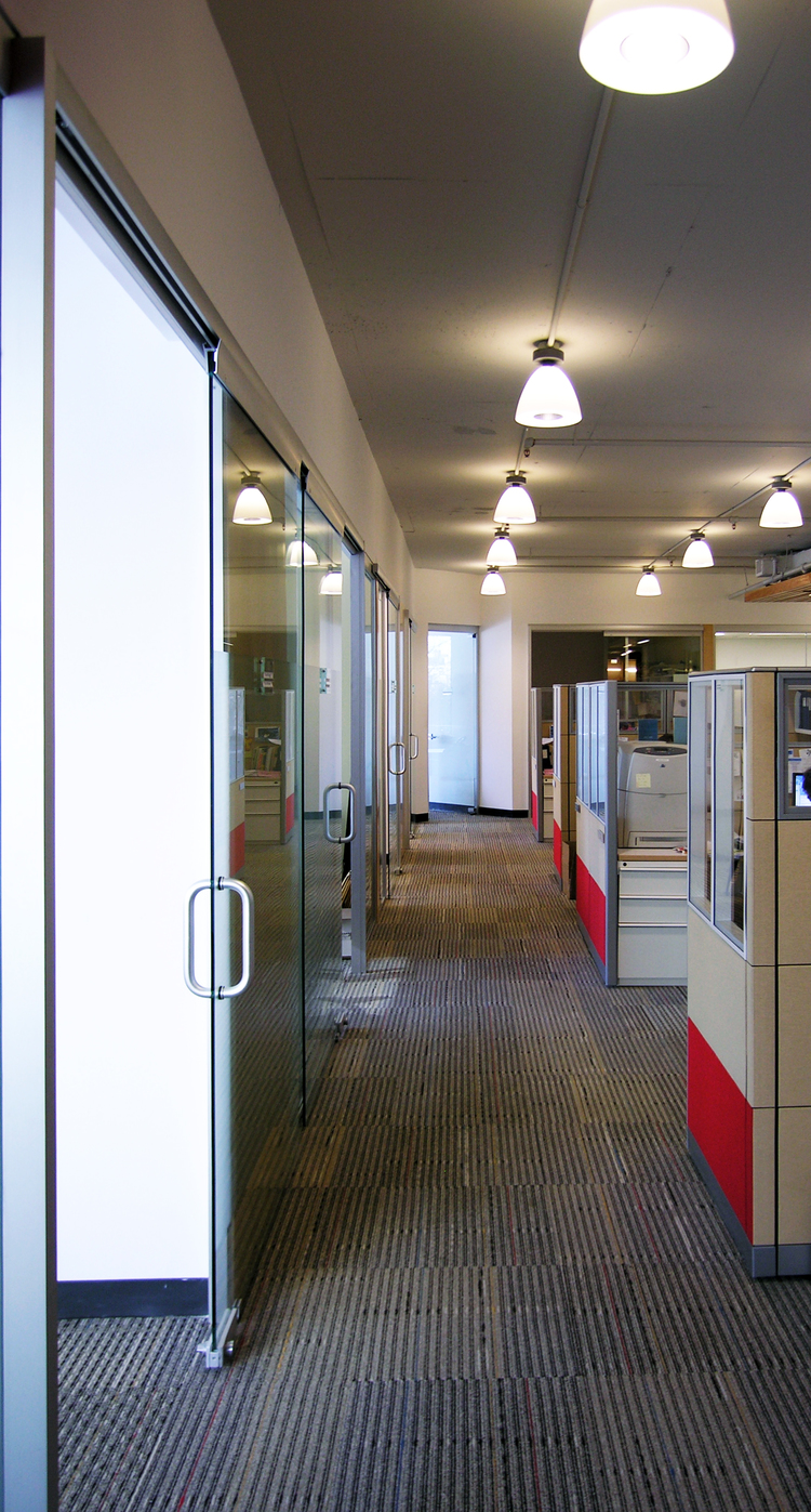 Open cubicle zone at Spence-Chapin with glass-enclosed private offices along the exterior wall, under a ceiling of pendant lights.