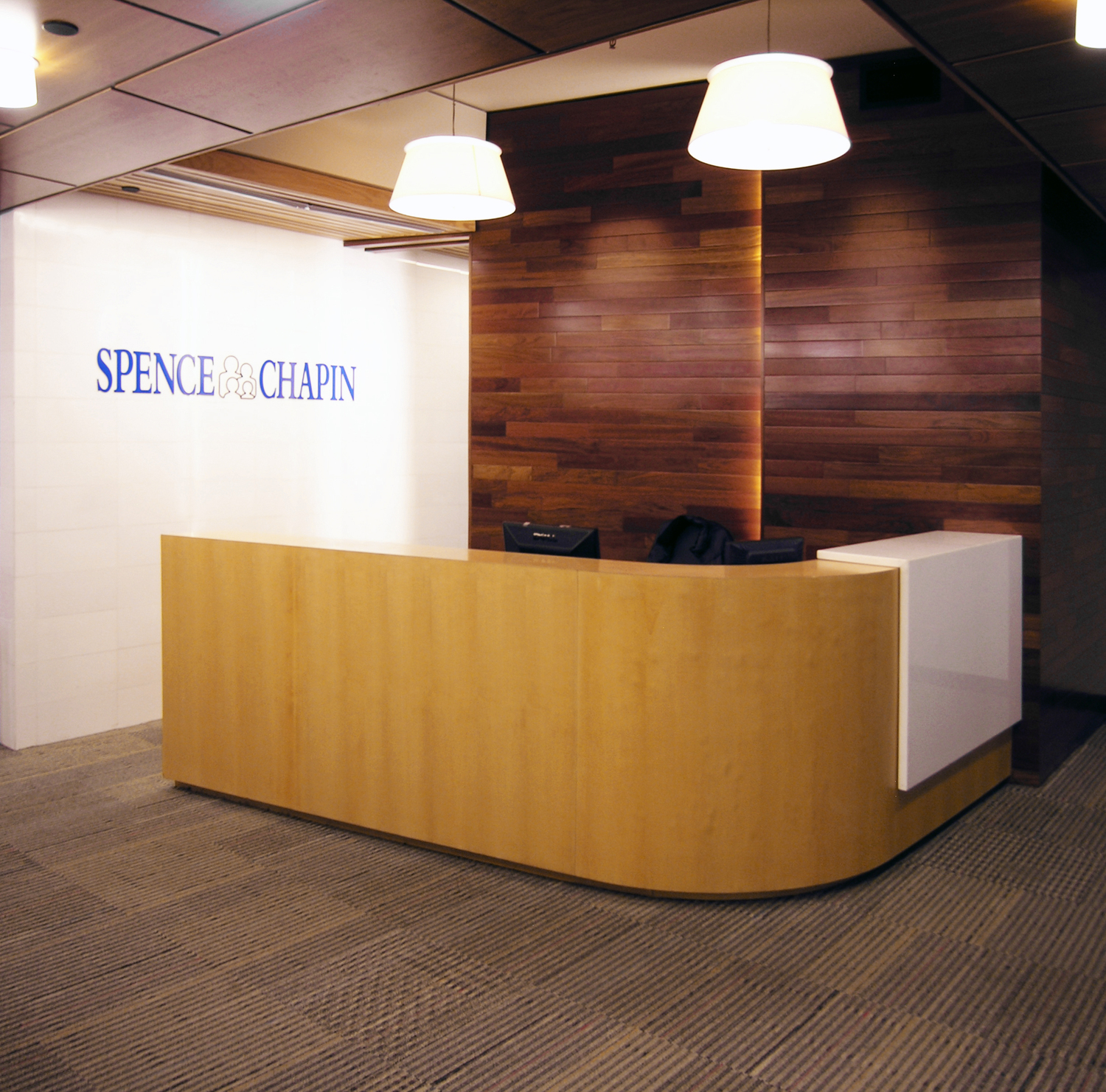 Spence-Chapin reception desk in light maple wood set in front of a dark horizontal wood wall, with the organization’s logo on an adjacent white wall and two pendant fixtures above.