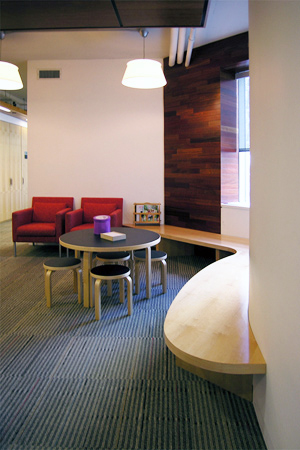 Children’s waiting area at Spence-Chapin with a low round play table, small stools, and two red lounge chairs set against a wood-paneled accent wall and curved built-in bench.