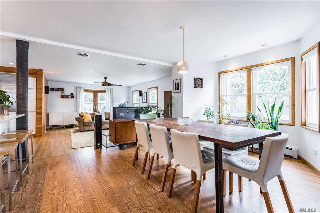 Large dining table at Walnut House surrounded by upholstered chairs, positioned beside corner windows with plants and open to the living area and kitchen.