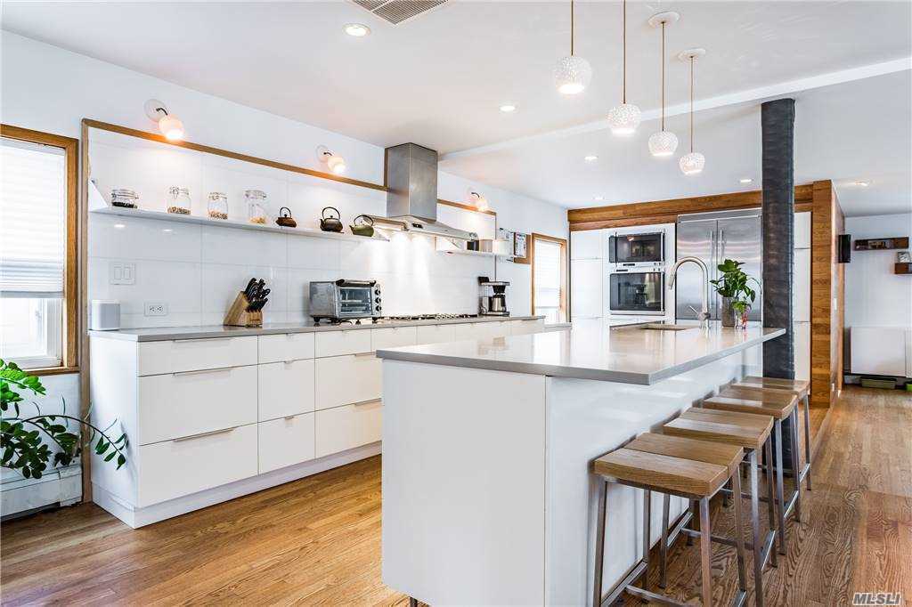 Open kitchen at Walnut House with long white island, counter stools, and a wall of white cabinets and shelving, all set on continuous wood floors.