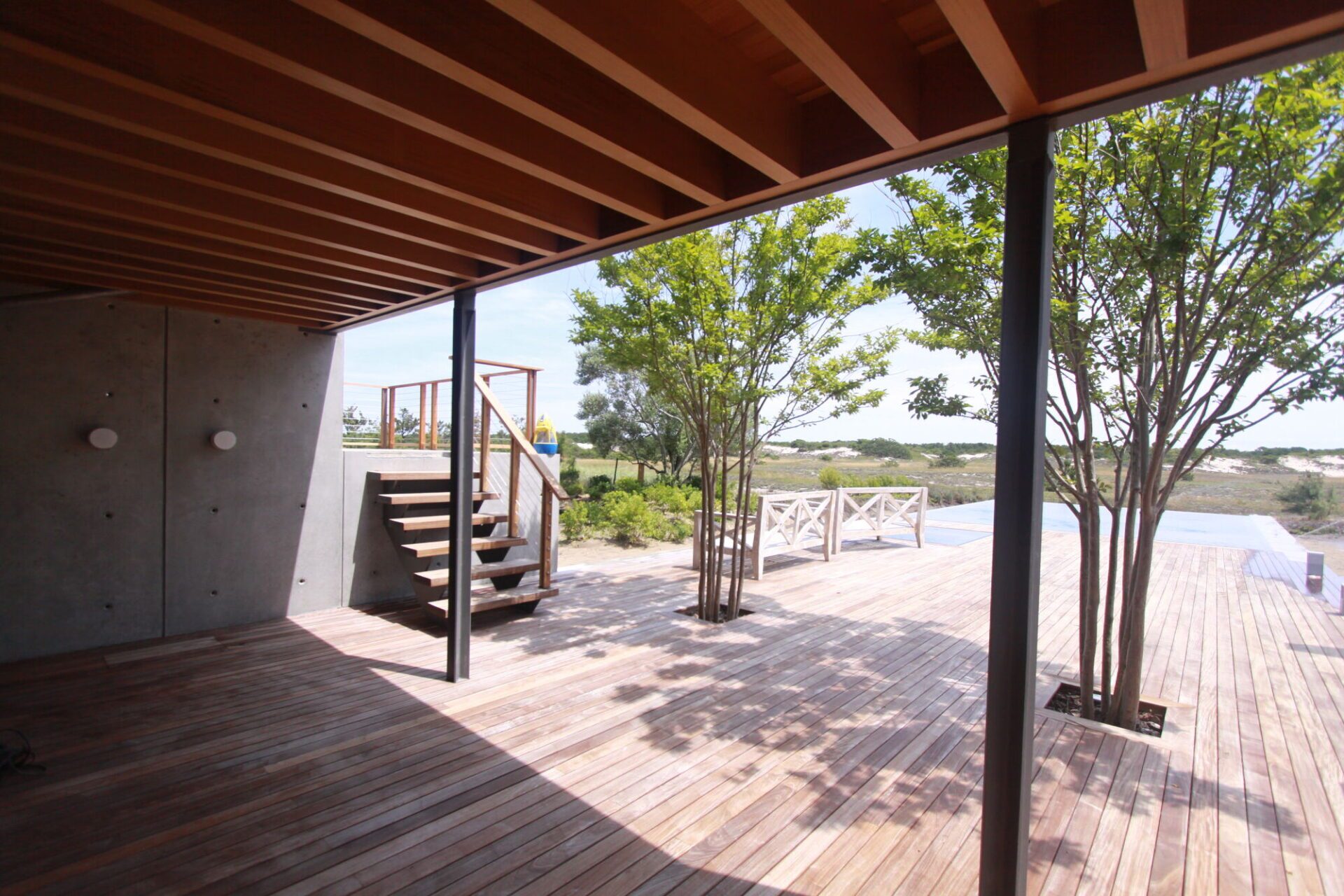 Covered ipe terrace under exposed wood beams, with a smooth concrete wall and reveals at left, wood steps, a steel column and the pool and dunes beyond.