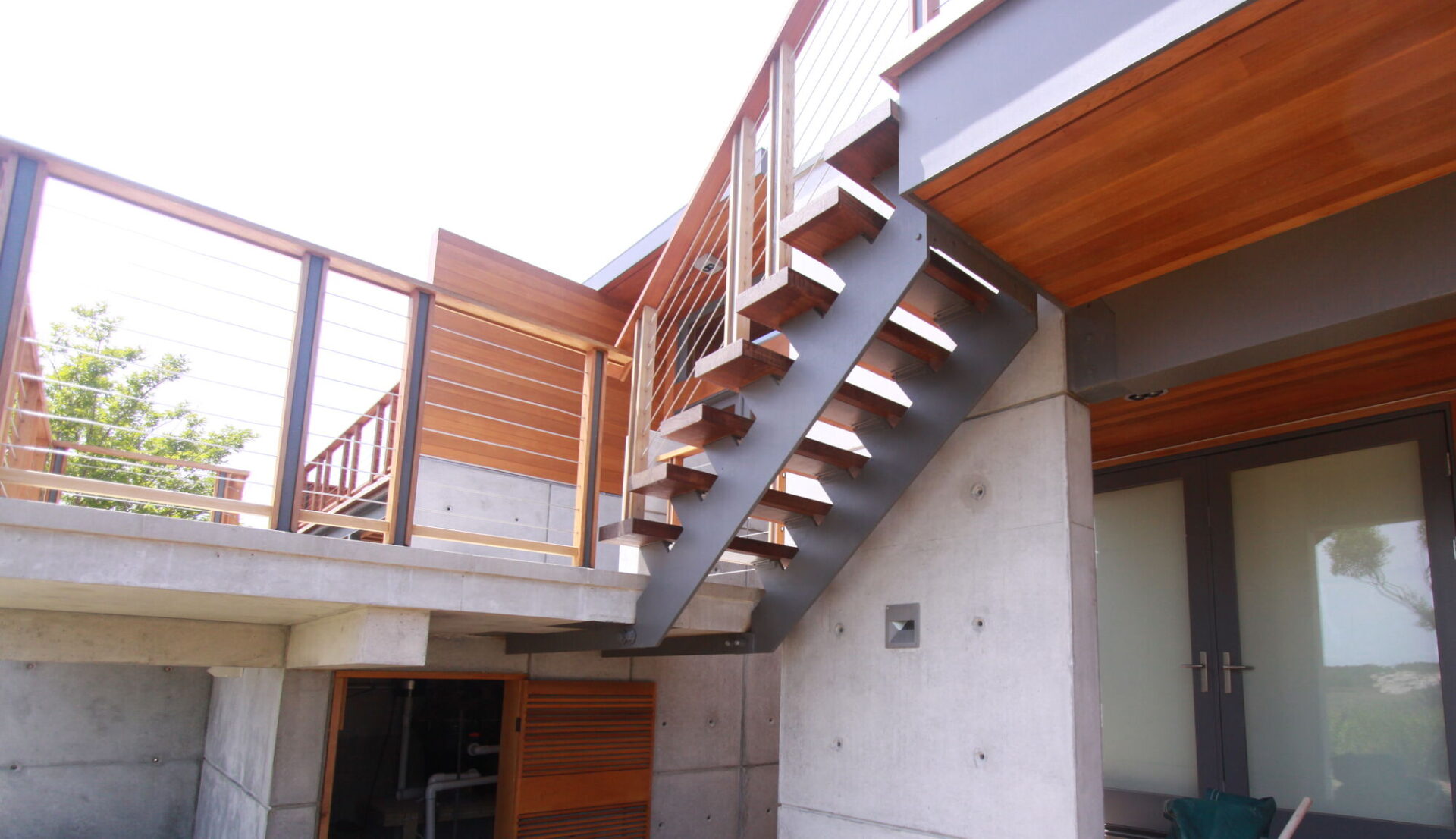 Exterior steel stair with ipe treads and a cable rail climbing beside a smooth concrete wall to an upper deck, with cedar siding and glass doors beyond.