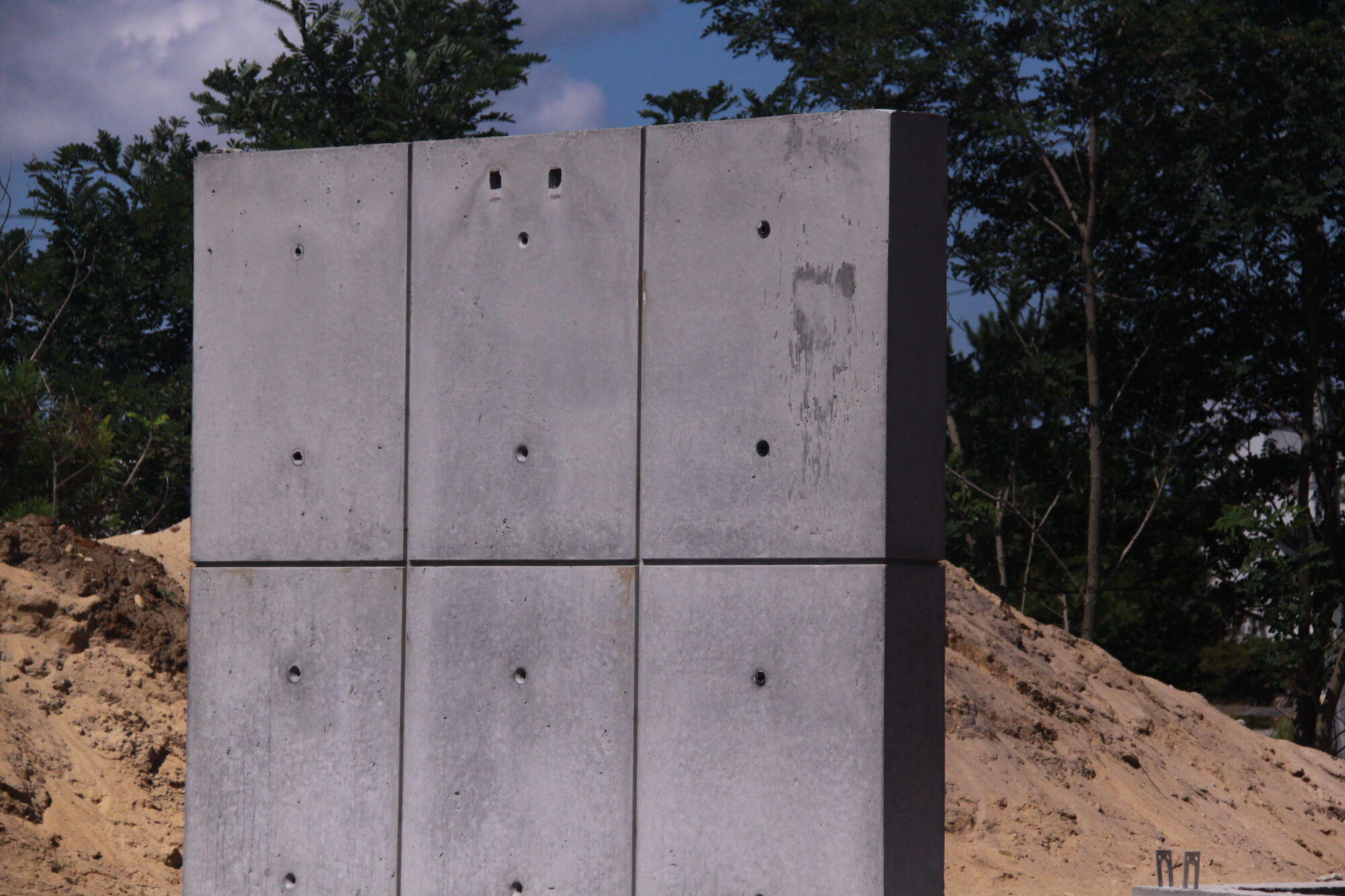 Freestanding concrete wall with a grid of reveals and tie holes, set against trees and a sand berm.