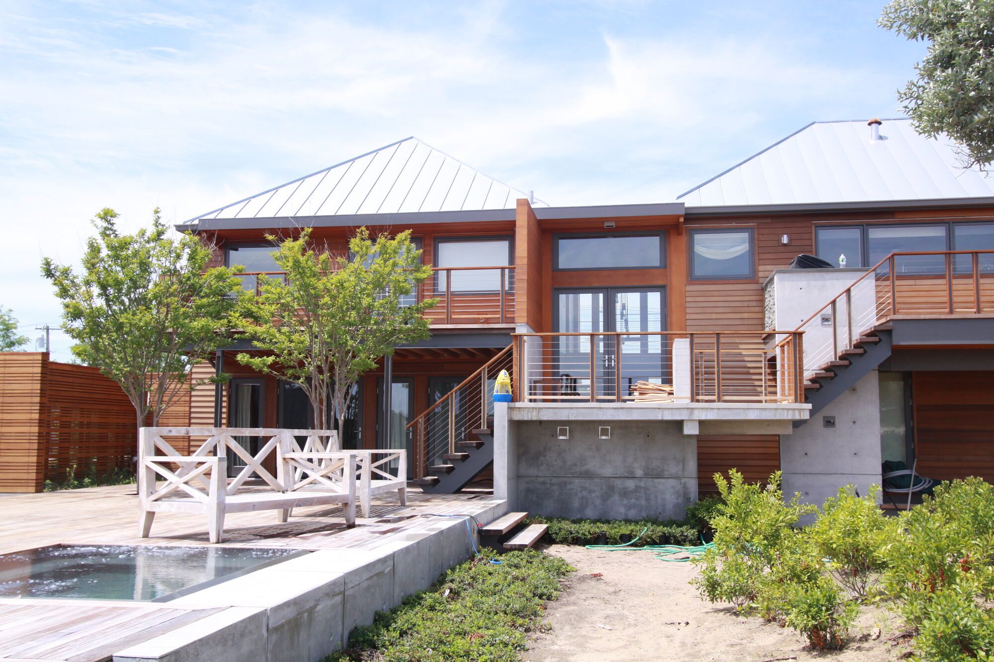 Rear of a cedar-clad two-story house with steel stairs, a concrete base wall and a concrete-edged spa and deck in the foreground.