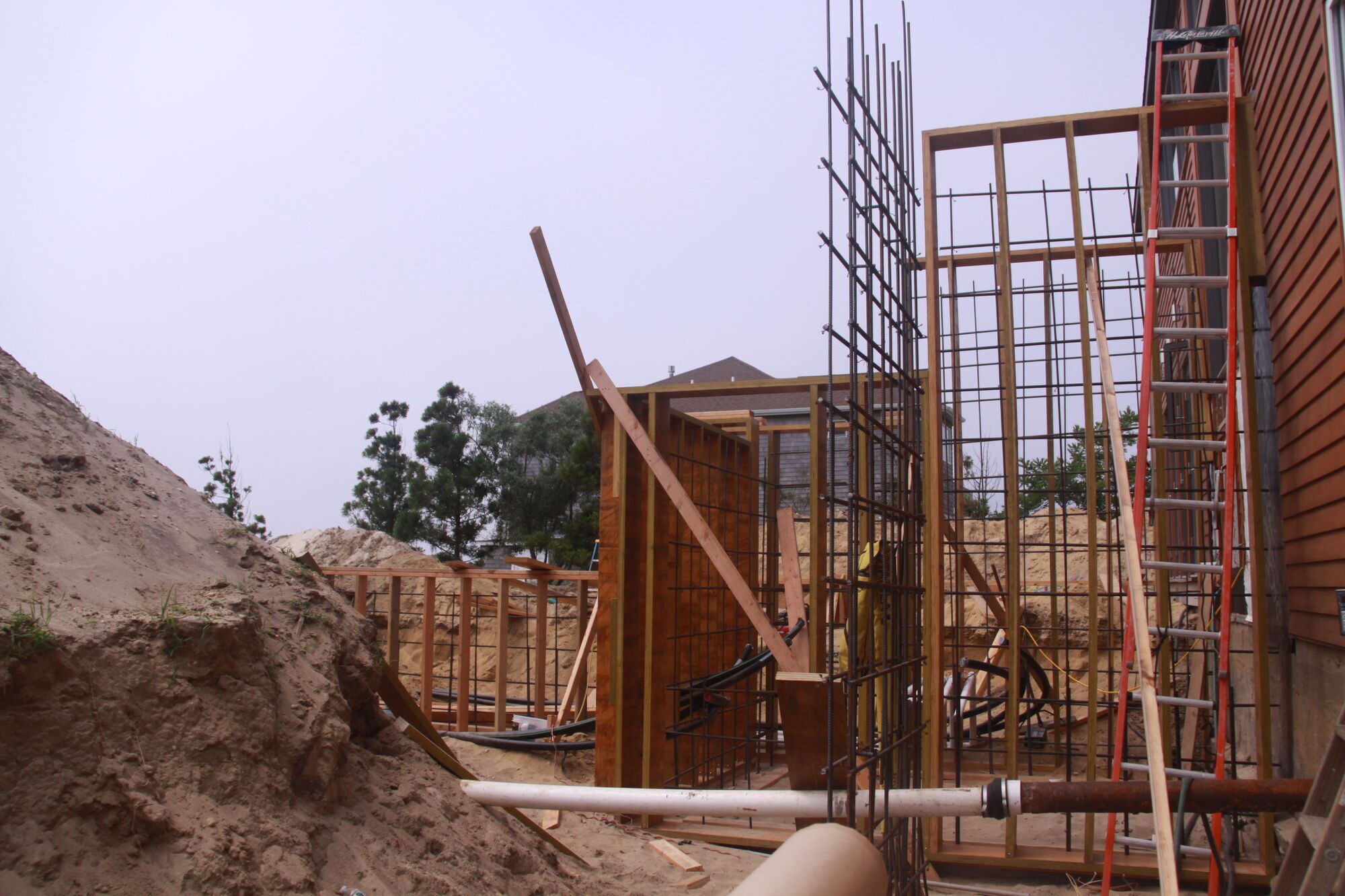 Vertical rebar cages and wood formwork rising next to a cedar-clad house on a sandy site.
