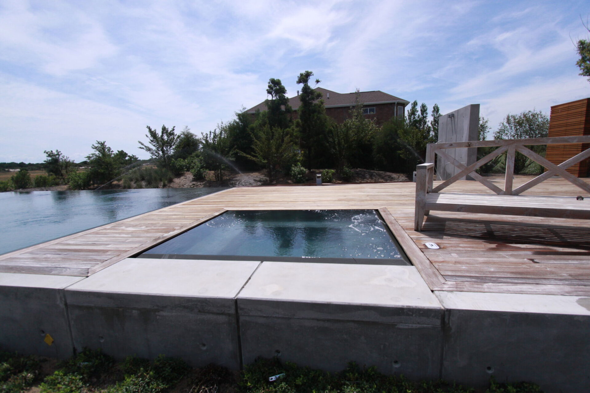 Square spa filled to the brim, set into a wood deck with a concrete base wall and the pool beyond.